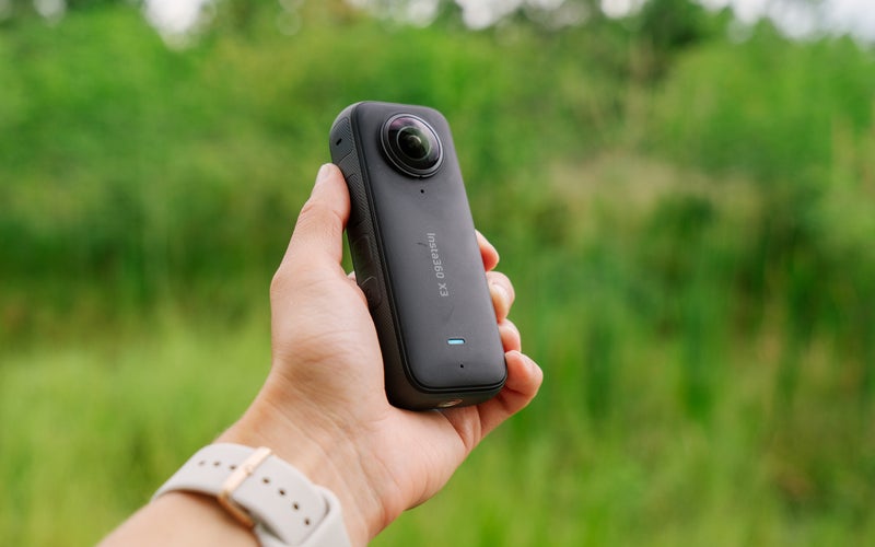The Insta360 X3 held in a hand in front of a green forest