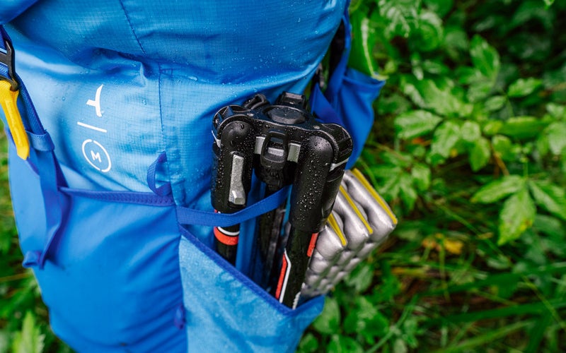 A black Manfrotto Befree Advanced carbon fiber tripod sits in the water bottle pocket of a blue hiking backpack.