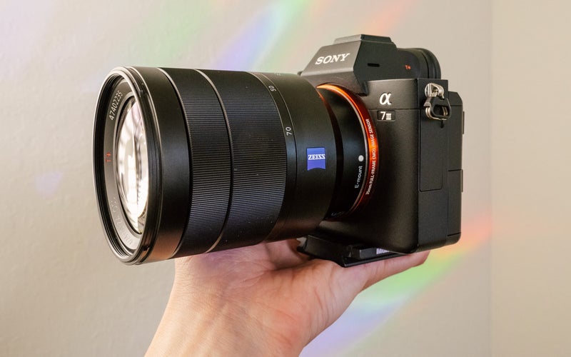A hand holds the Sony Vario-Tessar T* FE 24-70mm f/4 Lens and Sony a7 III camera in front of a white wall with rainbows.